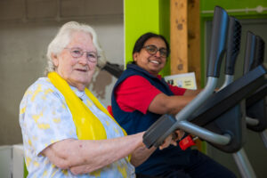  Two women are exercising on elliptical machines in a gym with bright green walls. The woman in the foreground has white hair and is wearing glasses, a light-colored patterned shirt, and a yellow scarf. She is smiling and looking towards the viewer. The woman behind her is also smiling and looking towards the viewer, wearing glasses, a red shirt, and a dark blue vest.
