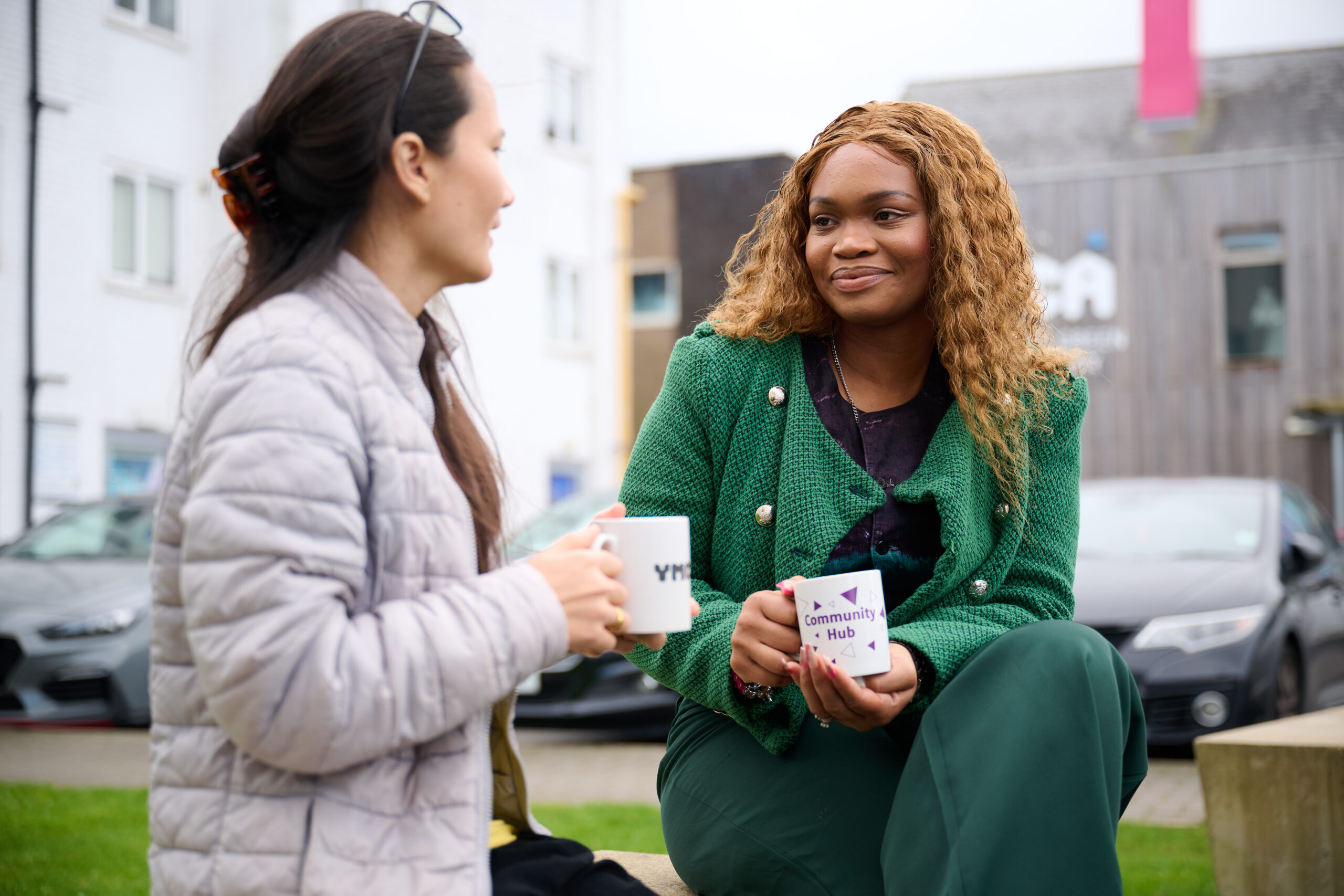  Two women are seated outdoors, holding mugs and engaging in conversation. The woman on the left, with dark hair pulled back, wears a light grey quilted jacket. The woman on the right has long, curly hair and is wearing a green textured jacket and dark green trousers, looking towards the other woman with a slight smile. There are buildings and cars in the background.